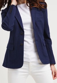 Blazer bleu marine en tissu texturé. Comprend deux boutons, deux poches avant et une poche poitrine. Porté par-dessus une chemise blanche.