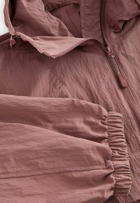 Giacca leggera rosa con cappuccio, dotata di zip frontale e polsini arricciati; tessuto texture con finitura stropicciata.