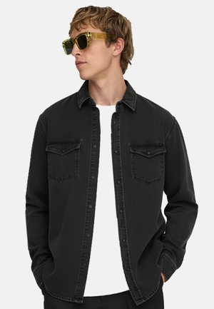 Mies, joka käyttää keltavihreitä aurinkolaseja, mustaa napitettavaa paitaa, jossa on rintataskut, valkoinen t-paita alla, katsoo vasemmalle, kädet taskuissa.