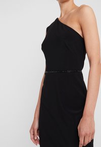 Svart enaxlad klänning med en figurnära silhuett, som har ett pärlbesatt midjeband och slät, stretchig material. Enkelt, elegant design.