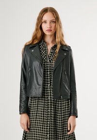 Jaqueta de motoqueiro em couro preta com bolsos com zíper, detalhes em prateado e um design curto, usada sobre um vestido xadrez preto e branco.