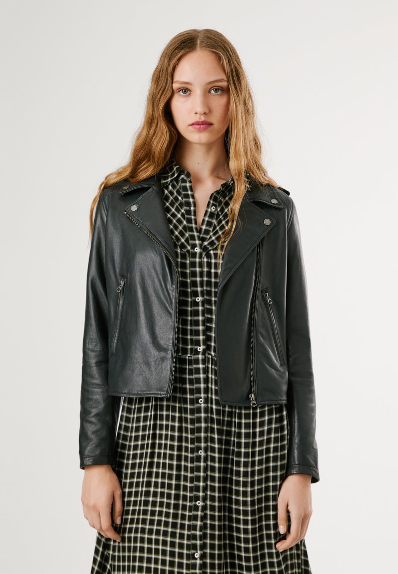 Jaqueta de motoqueiro em couro preta com bolsos com zíper, detalhes em prateado e um design curto, usada sobre um vestido xadrez preto e branco.