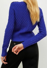Jersey de punto morado con un patrón de espiga, dobladillo acanalado y puños de mangas, con detalle de botones en el hombro. Se muestran pantalones negros.