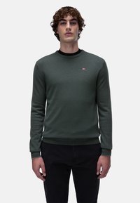 Maglione verde lavorato a maglia con scollo rotondo, polsini e orlo a coste, con un piccolo emblema. Abbinato a pantaloni neri.