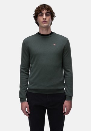 Groene gebreide trui met een ronde hals, geribde manchetten en zoom, met een klein embleem. Gecombineerd met zwarte broek.