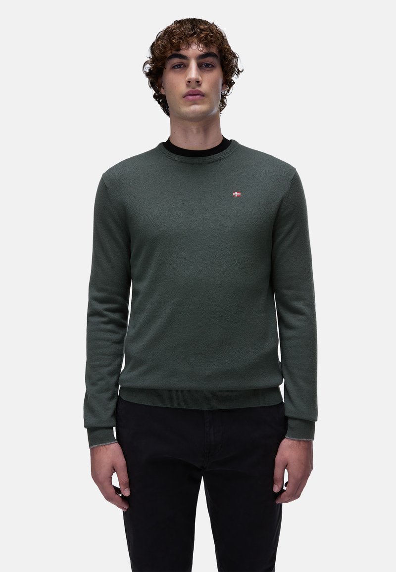 Maglione verde lavorato a maglia con scollo rotondo, polsini e orlo a coste, con un piccolo emblema. Abbinato a pantaloni neri.
