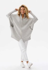 Jasnoszara, oversize sweter dzianinowy z zamkiem błyskawicznym, prążkowanymi rękawami i kieszeniami po bokach. Połączony z białymi spodniami i trampkami.