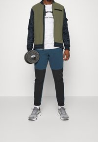 Pánský sportovní outfit sestávající z dvoubarevné bomber bundy v olivově zelené a černé barvě, bílého trička Under Armour a modro-černých teplákových kalhot, držící činku.