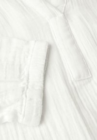 Tela de algodón blanca con una textura arrugada, que presenta una abertura con pliegue y un borde de manga doblado. Material suave y ligero.