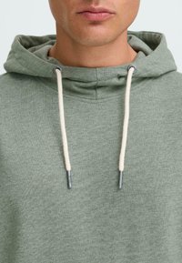 Sudadera verde de algodón con cordón y detalles de acero inoxidable. Textura suave y un corte casual, con un bolsillo canguro estándar.