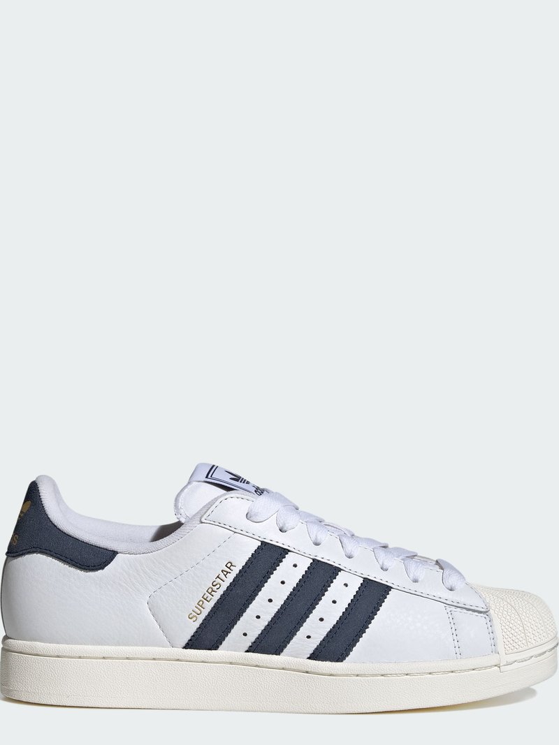 White Adidas Superstar sneaker with black stripes, gold "SUPERSTAR" text, and rubber shell toe, shown from the side on a plain background.