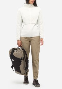 Osoba w białej polarowej kurtce i beżowych spodniach trzyma dużą beżowo-czarną plecak turystyczny za uchwyt.