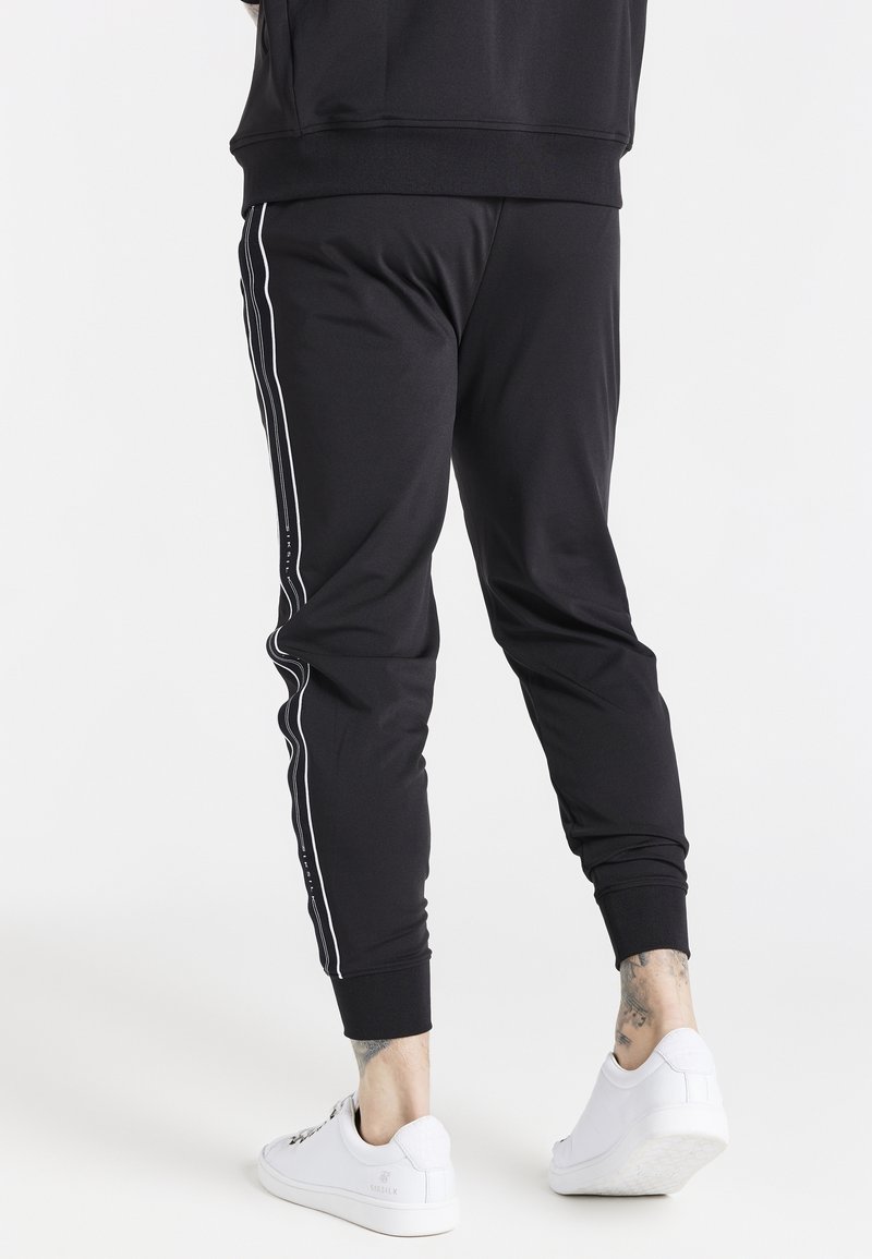 Black jogger pants made of stretchy fabric, featuring white side stripes, elastic cuffs, and a slim fit. Paired with white sneakers.