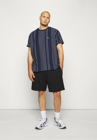 Camicia a righe navy a maniche corte con taschino, abbinata a pantaloni neri. Le scarpe sono bianche con dettagli navy. Design pulito e casual.