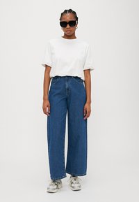 White cotton t-shirt, relaxed fit, paired with high-waisted, wide-leg blue denim jeans. Black rectangular sunglasses and patterned sneakers.