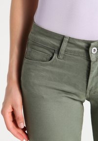 Pantalones de mezcla de algodón verde con un diseño ajustado, estilo de cinco bolsillos, costuras visibles y cierre de botón plateado. Textura suave.