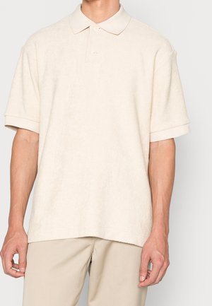 Cream-colored polo shirt made of soft terry fabric, featuring short sleeves, a collar, and two buttons at the neckline.