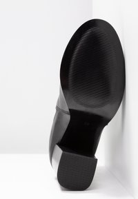 Semelle de chaussure en cuir noir avec une accroche texturée et un talon bloc, positionnée en angle contre une surface blanche d’angle, affichant la taille 37.
