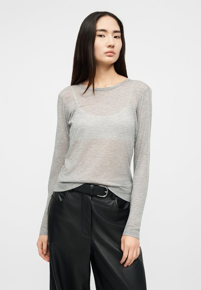 Žena s dlhými tmavými vlasmi, oblečená v priehľadnom sivom tričku s dlhým rukávom a čiernych kožených nohaviciach s čiernym opaskom, stojaca pred jednoduchým pozadím.