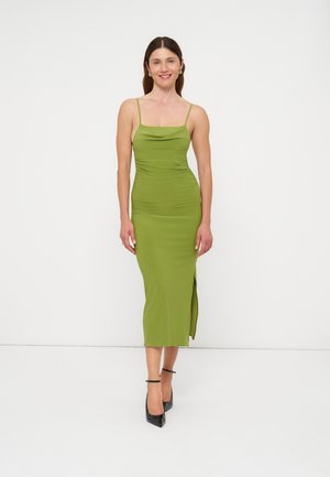 Femme souriante, portant une robe midi verte ajustée sans manches avec fines bretelles et fente sur le côté, talons noirs, debout sur un fond blanc uni.