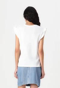 T-shirt blanc en coton avec des manches volantées, associé à une jupe en denim bleu. Le tissu semble doux avec une texture lisse.