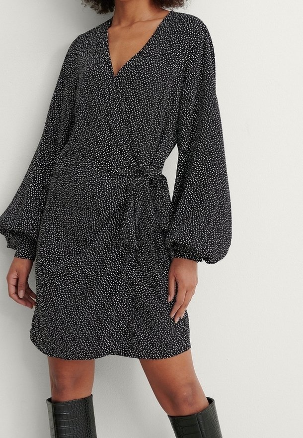 Femme portant une robe portefeuille noire à pois blancs, avec de longues manches bouffantes et des bottes noires texturées jusqu'aux genoux.