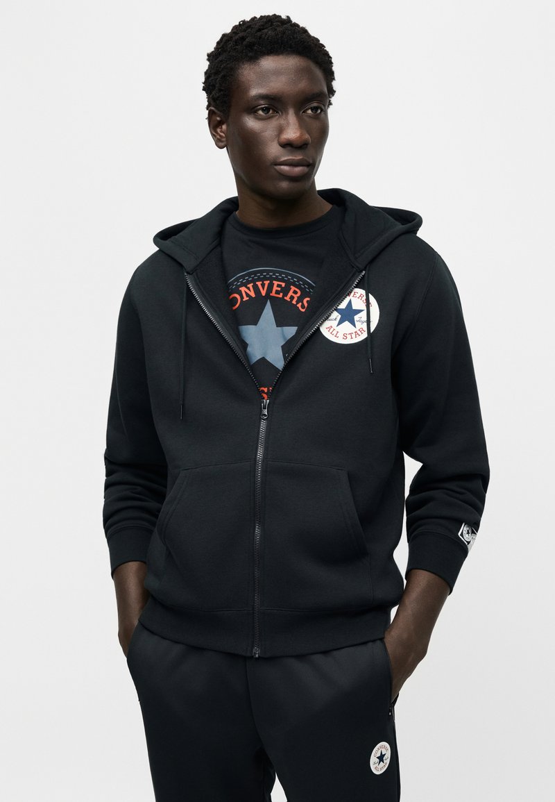 Sort lynlås-hoodie med justerbar hætte, forlommer og broderet logo. Båret over en sort t-shirt med et cirkulært grafisk design.