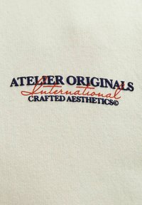 Krémporszínű pamut falikép, hímzett felirattal: "ATELIER ORIGINALS International CRAFTED AESTHETICS©" sötétkék és piros színben.