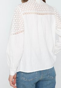 Biała bluzka wykonana z teksturowanej tkaniny z koronkowymi detalami na ramionach i plecach, wykończona mankietami na guziki oraz prostym dołem.