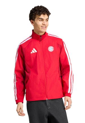 Junger Mann lächelt und schaut nach rechts, trägt eine rote FC Bayern München Zip-Up Jacke mit weißen Adidas-Streifen an den Ärmeln.