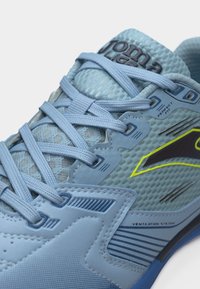 Αθλητικό παπούτσι ανοιχτού γαλάζιου χρώματος με πλέγμα στο πάνω μέρος, λεπτομερή κορδόνια, επωνυμία "Joma" και κίτρινη λεπτομέρεια στο πλάι, με ετικέτες για εξαερισμό και τέλεια εφαρμογή.