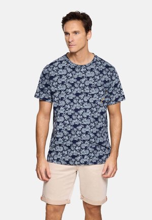 Hombre con camisa de manga corta de estampado floral azul marino y pantalones cortos beige remangados, de pie contra un fondo claro y liso.