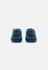 Kék velúr sportcipők fekete részletekkel, sárga díszítéssel az oldalon, valamint texturált sarokkal, amelyen kék Nike logó található.