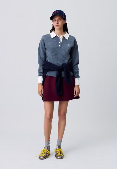 Suéter gris de punto con un collar blanco, falda burdeos y suéter azul marino atado a la cintura. Zapatillas amarillas y grises con calcetines hasta el tobillo.