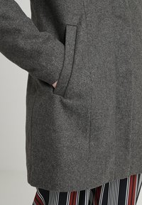 Abrigo de lana gris con un diseño limpio, bolsillos laterales y una textura suave. Se pueden observar rayas verticales en la tela debajo.