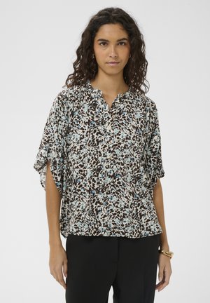 Göndör hajú nő laza, rövid ujjú blúzt visel absztrakt fekete, fehér és kék mintával, fekete nadrággal és arany karkötővel párosítva.