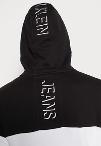 Schwarz-weißer Kapuzenpullover mit einem auffälligen "JEANS"-Grafikdruck auf dem Rücken, weichem Stoff und kontrastierendem Farbschema.