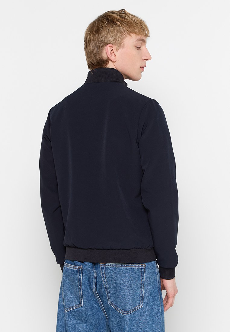 Chaqueta de bombardero azul marino con un alto cuello, puños y dobladillo acanalados. Tela suave con un corte relajado, combinada con jeans de mezclilla azul.