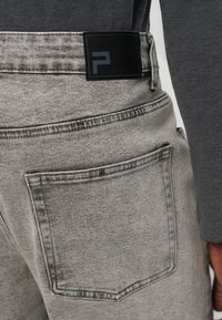 Κοντινό πλάνο τσέπης από τζιν γκρι ανοιχτό και μαύρη ορθογώνια ετικέτα με ένα μινιμαλιστικό γεωμετρικό λογότυπο στη μέση, φορεμένη με σκούρο γκρι τοπ.