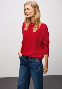 Roter, lockerer Pullover mit langen Ärmeln und weitem Ausschnitt, kombiniert mit blauen Jeans. Glatte Textur, lässiger Stil.