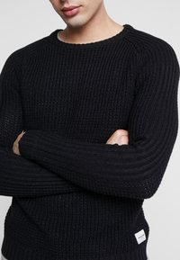 Czarny sweter dzianinowy z teksturowanym wzorem prążkowym, okrągłym dekoltem i dopasowanymi rękawami. Posiada małą metkę z logo na dolnej stronie.