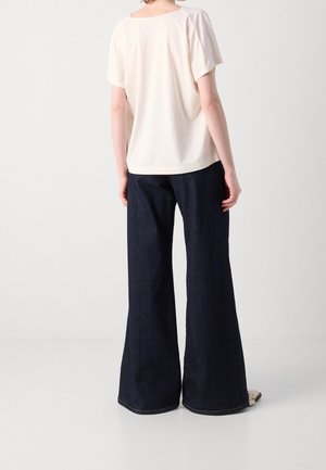 Personne portant un haut blanc ample à manches courtes et un jean en denim sombre à jambes larges, debout devant un fond clair uni.