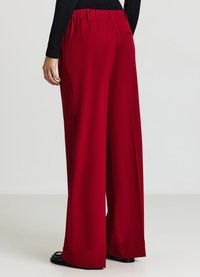 Pantaloni a gamba larga in un ricco tessuto rosso. Vita elastica, texture liscia e orli arrotolati. Presenta due tasche laterali per la funzionalità.