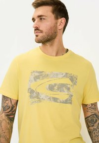 Gul bomulds T-shirt med et gråt grafikdesign med en struktureret finish. Korte ærmer, rund halsudskæring og synlig tatovering på armen.