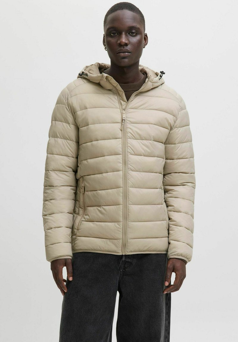 Bežs pufīgs/ puffer/ jakas ar kapuci, kas aprīkota ar horizontālu quilted/ siltinājuma dizainu, ar rāvējslēdzēju un divām sānu kabatām, uzvilkta virs brūnas džempera.