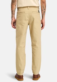 Pantalones de algodón beige con corte recto, dos bolsillos traseros y una etiqueta en la cinturilla, combinados con zapatos de cuero marrón.