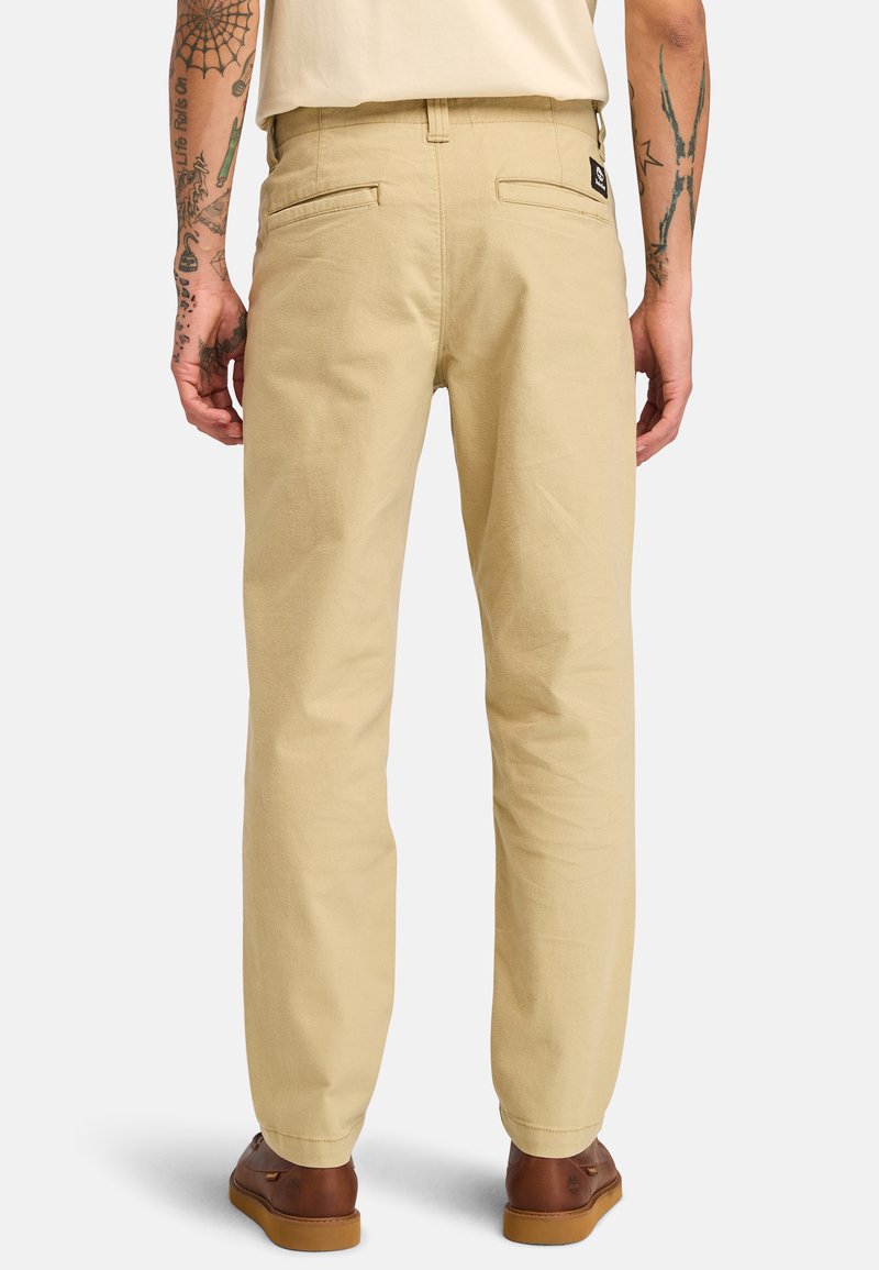 Pantaloni in cotone beige a taglio dritto, con due tasche posteriori e una etichetta sulla cintura, abbinati a scarpe in pelle marroni.