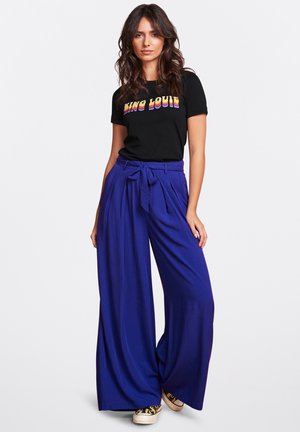T-shirt noir avec un texte coloré "KING LOUIS", associé à un pantalon large violet taille haute avec une ceinture nouée.