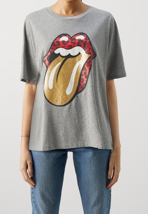 Persona vistiendo una camiseta gris con un gran gráfico de labios de leopardo rojo y lengua dorada, combinada con jeans azules.