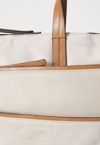 Sac en toile beige avec des poignées et des finitions en cuir marron, doté d'une surface texturée et de coutures visibles le long des bords.